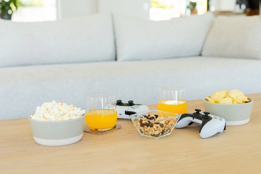 Wooden coffee table near sofa holding popcorn and chip bowls, nut dish, orange juice, controllers