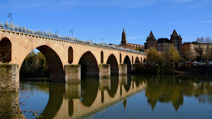 Fototapeta premium Scenic View of Historic Stone Bridge Spanning Over Serene River with Cityscape Background Under Clear Blue Sky in Montauban Setting