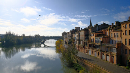 Fototapeta premium Montauban Scenic view of a tranquil river reflecting the clear blue sky, lined by historic buildings with charming architecture and a distant bridge under fluffy clouds