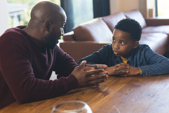 African American father and son sitting at wooden table in living room, gesturing toward glass bowl