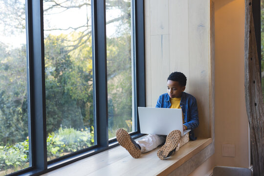 African American boy child sitting on wooden bench studying on laptop by grid window, copy space