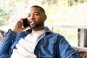 African American man sitting on leather sofa at home under large window talking on smartphone