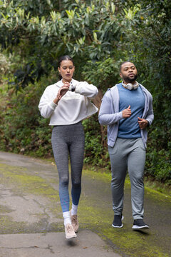Diverse friends jogging along narrow wooded park trail checking smartwatch and wearing headphones