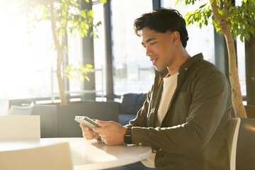 Asian man interacting with smartphone while sitting at table by glass windows in cafe, copy space