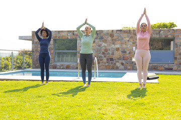 Diverse female friends practicing yoga on backyard lawn by pool with rolled mats and pool ladder