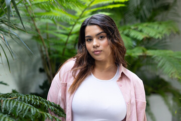 Woman standing among tropical ferns and palms at greenhouse, wearing white tank top and pink shirt