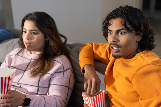Couple sitting on sofa at home holding striped popcorn cartons while watching TV with surprise