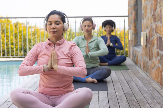Diverse women sitting on pool deck meditating on yoga mats in activewear and over-ear headphones