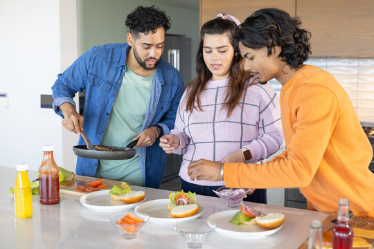 Diverse friends standing around kitchen island cooking burger patties in pan, adding onion slices - Powered by Adobe