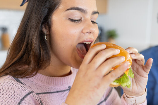 Woman holding hamburger close to mouth while sitting at kitchen counter in pink geometric sweater