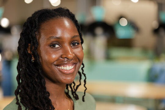 African adult woman smiling at camera in cafe with pendant lamps, wearing green top and hoops