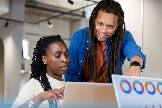 African American coworkers leaning at office desk analyzing pie charts on 2 laptops with smartwatch
