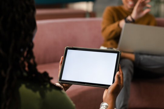 Tablet is being held on pink sofa in lounge, displaying blank white screen with blurred laptop