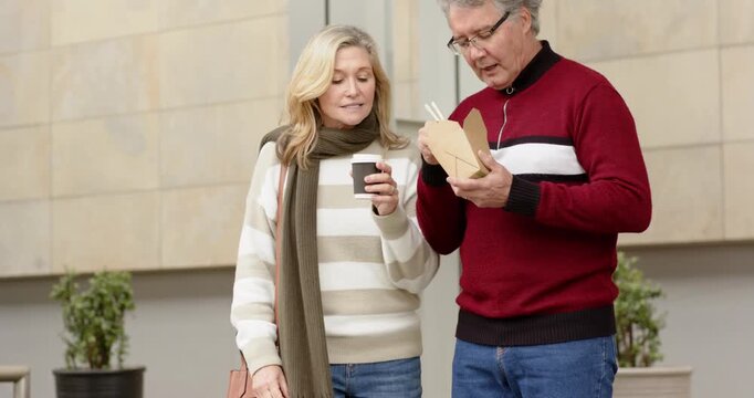 Senior couple lifting noodles from container on urban sidewalk during break, sipping coffee