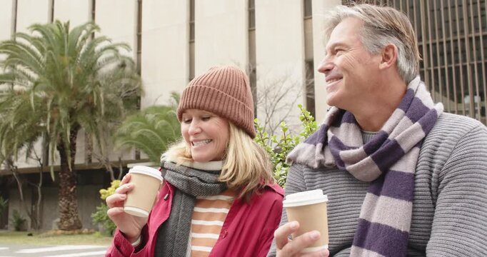 Senior couple holding coffee cups on sidewalk, woman tasting to test flavor, man gesturing laughing