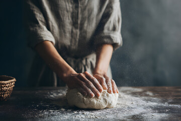 Fototapeta premium Close-up of female hands working with dough on a wooden surface, linen clothing and dark background highlighting the intimate, traditional bread-making moment