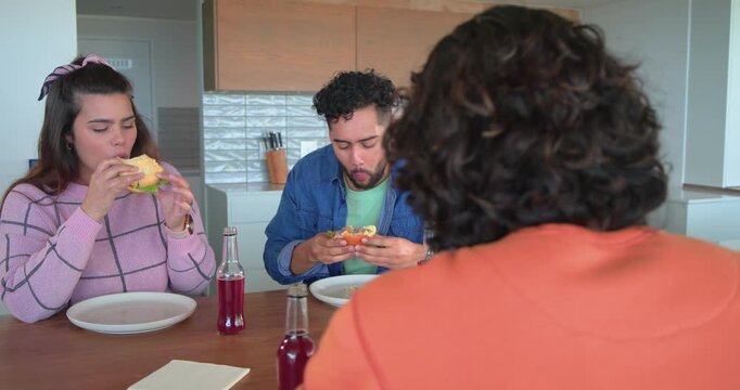 Friends taking bites of burgers at wooden kitchen island, chewing and discussing taste differences