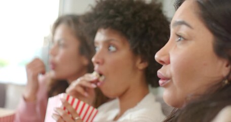 Women friends watching offscreen while eating popcorn tubs on sofa and central woman showing shock - Powered by Adobe