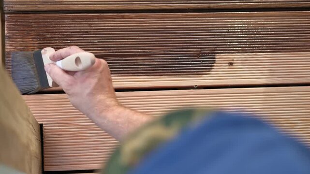 Brown oil for protection of wood by staining. Painting a larch for protecting and pests. Woodworker applying finish to terrace deck board. Repairman varnishing with brush wooden plank floor surface.