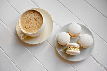 Top-down view of a white cup of frothy coffee with four white macarons on a small plate, a simple, elegant breakfast or dessert setting on a wooden table, minimal and inviting