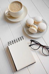 White wooden table displaying a cup of coffee, sweet macarons, a pair of reading glasses, and an open spiral notebook, perfect for representing planning, breakfast, or a refreshing work break