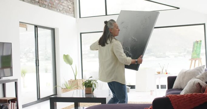 Mature adult White woman lifting canvas at home, pressing it onto wall from sofa, hanging art