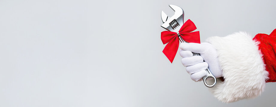 Santa's gloved hands holding a wrench with a red bow on a light gray background with space for text. Repair shop, auto mechanic. New Year in transportation