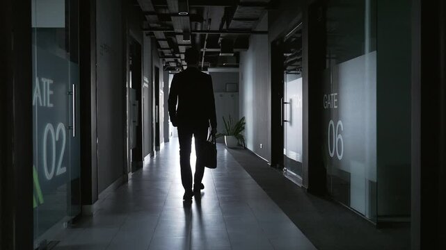 Silhouette of businessman in business suit walking down office corridor after work and meetings with briefcase in his hand. Rare back view man employee from behind towards the exit of the building