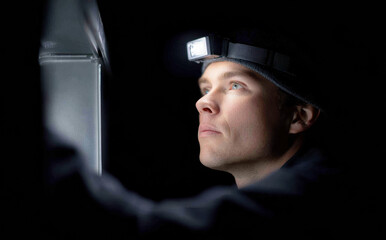Focused caucasian technician inspecting metal surface with headlamp in darkness expressing serious concentration and curiosity while working carefully at night