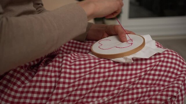 Woman's hands using a needle and red thread to create a heart pattern on white fabric held in a wooden embroidery hoop. A relaxing and creative handmade craft activity enjoyed at home