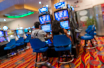 Fototapeta premium Blurred scene of people playing slot machines in a colorful casino with bright screens, blue chairs, and patterned carpet.