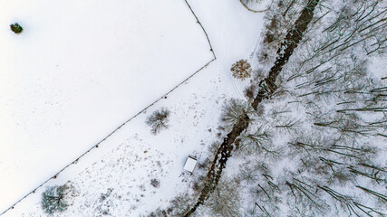 Aerial view of snowy field, fence line, and winter forest with a stream running through the landscape