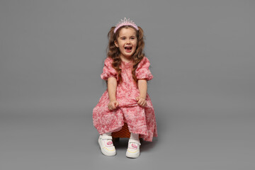Happy little girl in headband and dress sitting on grey background