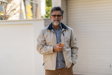 Smiling senior man outdoors holding a smartphone, dressed casually and looking confident in a bright, relaxed setting