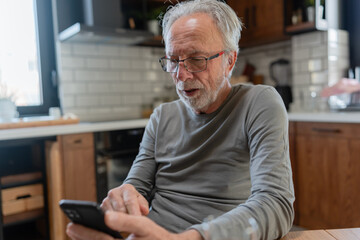 Elderly man sitting at a kitchen table, focused on his smartphone, with natural light illuminating the cozy modern kitchen behind him