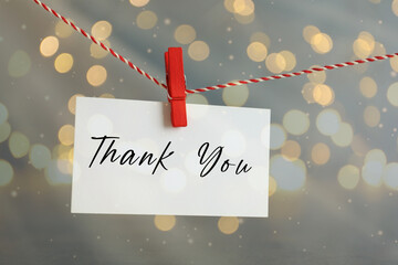 Thank You card hanging on thread against grey background with bokeh lights, closeup