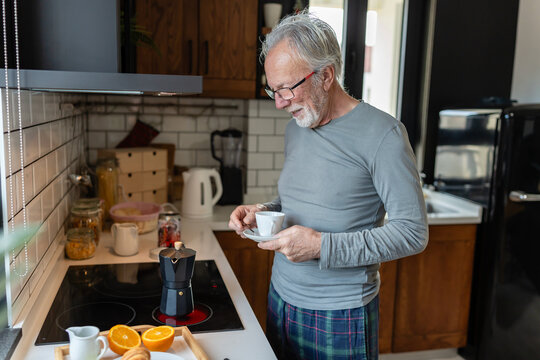 Smiling senior man preparing morning coffee in a cozy kitchen, holding a cup while waiting for a moka pot to brew beside fresh oranges - Powered by Adobe