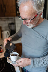 Elderly man pouring fresh coffee from a stovetop maker into a cup while standing in a cozy kitchen