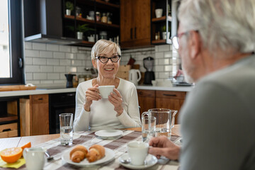 Smiling senior woman enjoying coffee at the breakfast table with her partner in a cozy kitchen setting