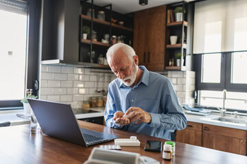 Senior man sitting in the kitchen, opening a pill bottle while using a laptop, managing medication and health tasks at home