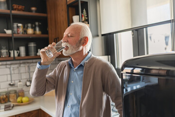 Senior man drinking a glass of water in a bright kitchen, taking a moment for hydration and daily...