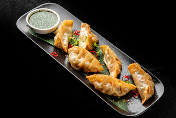 Golden pan-fried gyoza dumplings on a dark platter with green dipping sauce, chili, and herbs, captured in a dramatic, top-down perspective
