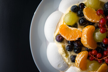 Close-up of a Pavlova meringue dessert topped with creamy filling and fresh berries, including red currants, blueberries and citrus slices. Bright directional light enhances textures and colors.