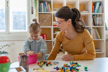 Fototapeta premium Caregiver and young girl sitting at a table, sorting colorful building blocks together in a bright and organized playroom