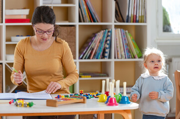 Young woman working at a table with colorful educational toys and papers, focused on planning or teaching activities in a bright classroom or home learning environment