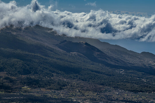 Vista de la cresta volc&aacute;nica de Cumbre Vieja con su nuevo volc&aacute;n de La Palma y el denso mar de nubes que corona la monta&ntilde;a. La ladera inferior muestra asentamientos y cultivos.