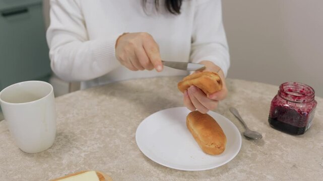 Mature woman wearing white knit sweater prepares sandwich using knife to spread berry jam from glass jar onto bread roll at beige marble table.