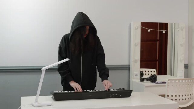 Musician wearing black hooded sweatshirt stands playing MIDI keyboard controller on white desk with white lamp and white basket in bright modern room with gray walls.