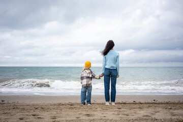 Mother and child standing hand in hand on the sandy beach, facing the ocean waves and cloudy sky, enjoying peaceful family time together by the sea