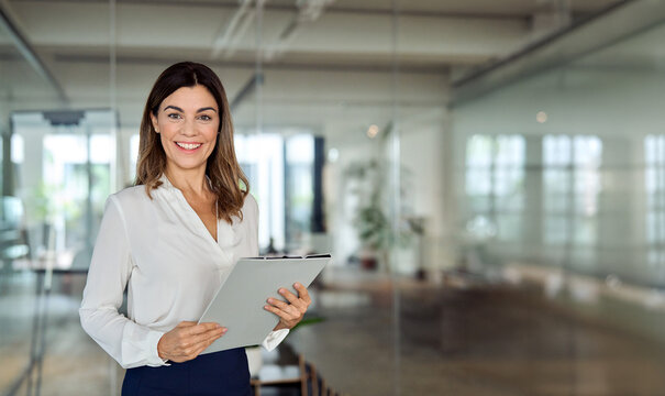 Happy 45 years old Latin professional middle aged business woman corporate leader, happy mature female executive, lady manager standing in office holding clipboard looking at camera, portrait. Banner